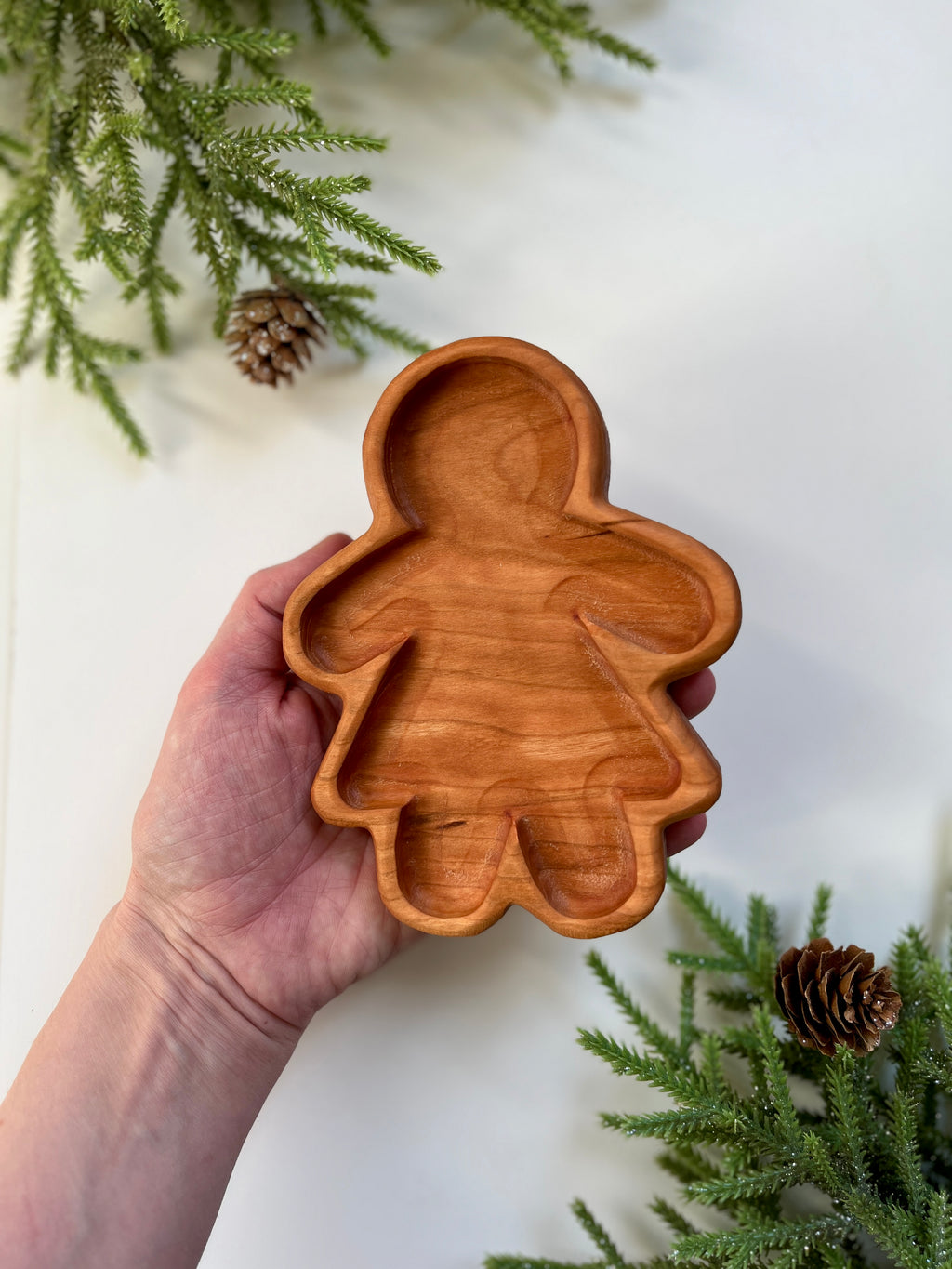 Hand holding a carved wooden tray shaped like a girl figure surrounded by pine branches and pine cones on white surface