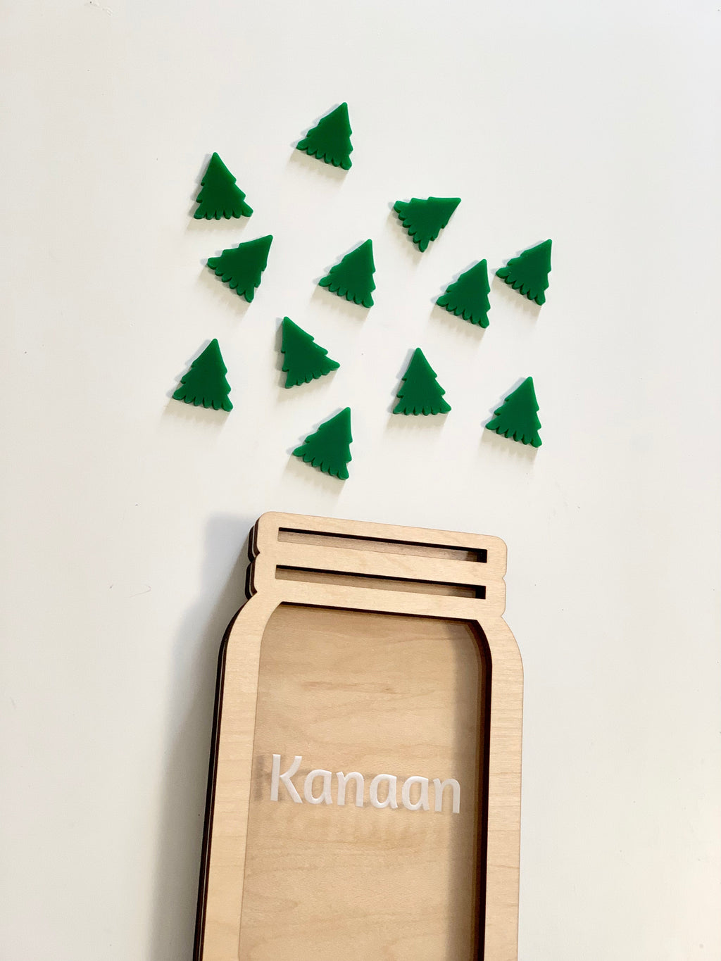 Light wooden jar-shaped frame with the name Kanaan and scattered green Christmas tree tokens on a white surface