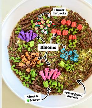 Bowl containing colorful flower-shaped flatbacks, vibrant blooms, green vines, leaves, and spring grass with dirt mix