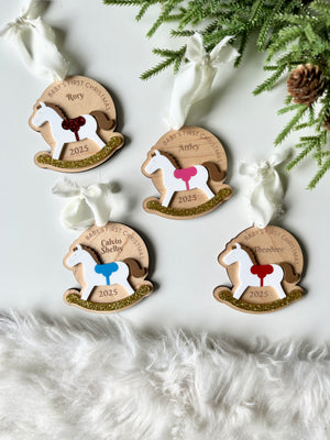Four personalized wooden rocking horse ornaments with colorful saddles and gold glitter bases on white background with greenery
