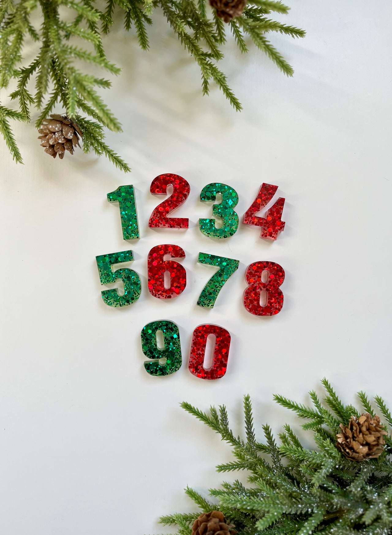 Green and red glitter resin numbers arranged on white surface with pine branches and pine cones framing the corners