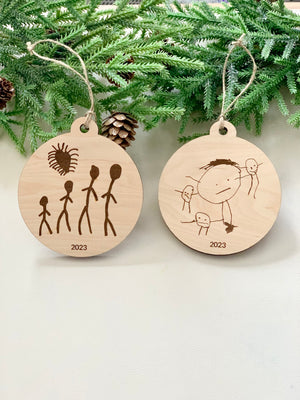 Two round wooden Christmas ornaments with childlike drawings and "2023" engraved, hanging with twine on pine branches