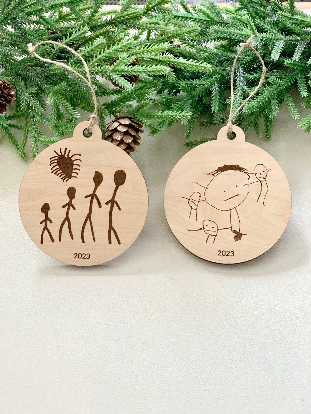 Two round wooden Christmas ornaments with childlike drawings and "2023" engraved, hanging with twine on pine branches