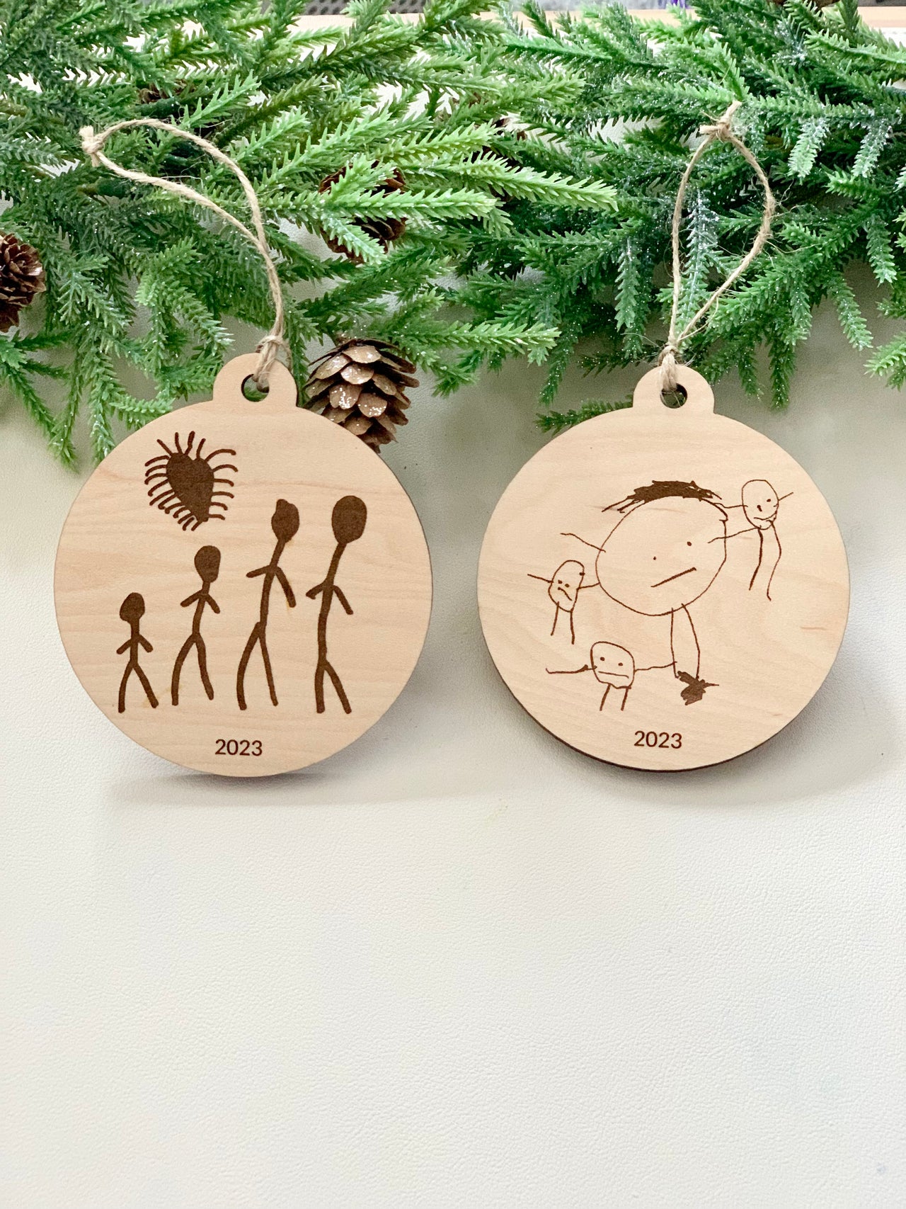 Two round wooden Christmas ornaments with childlike drawings and "2023" engraved, hanging with twine on pine branches