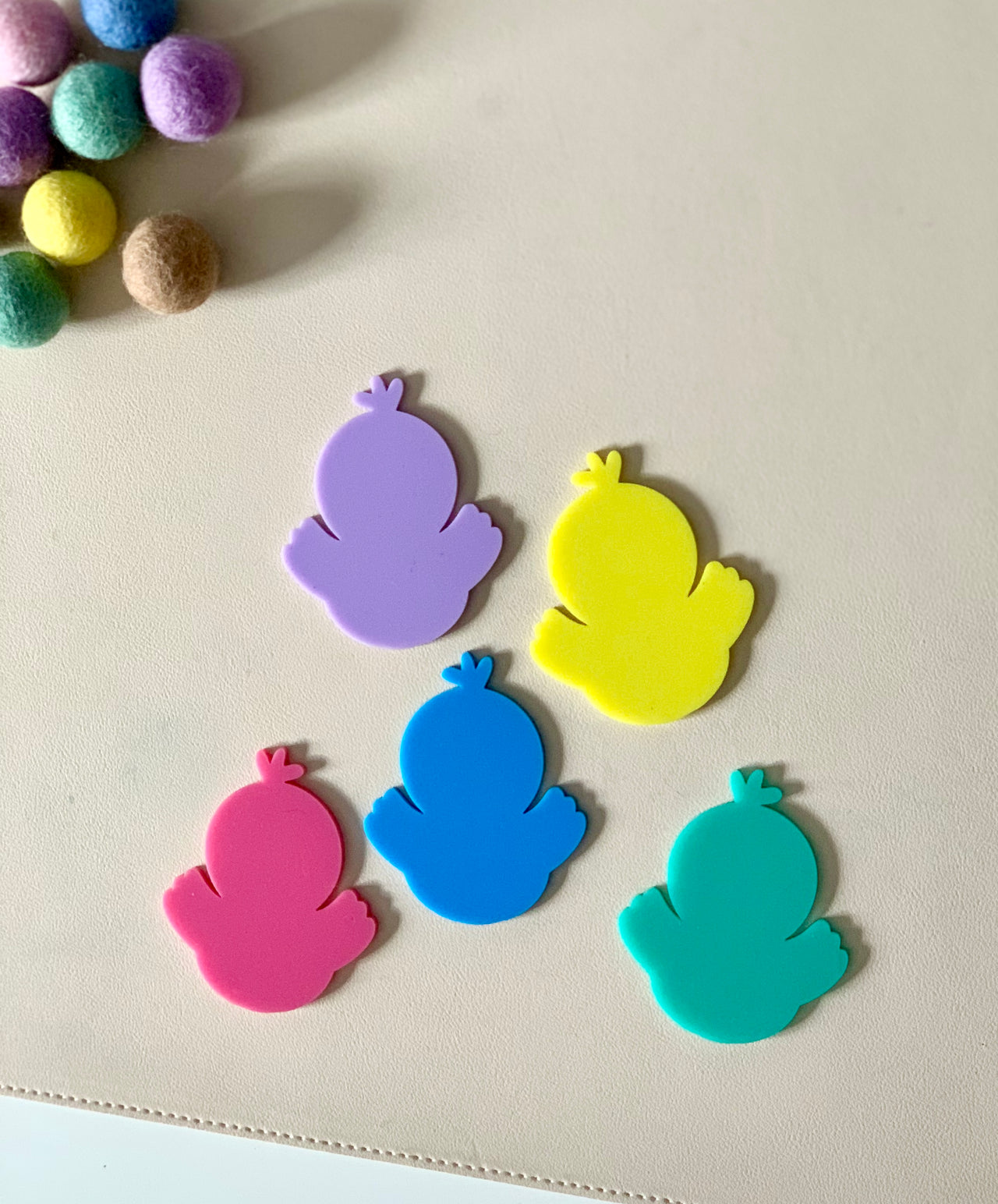 Five colorful chick-shaped cutouts in purple, yellow, pink, blue, and teal on a light surface near felt balls
