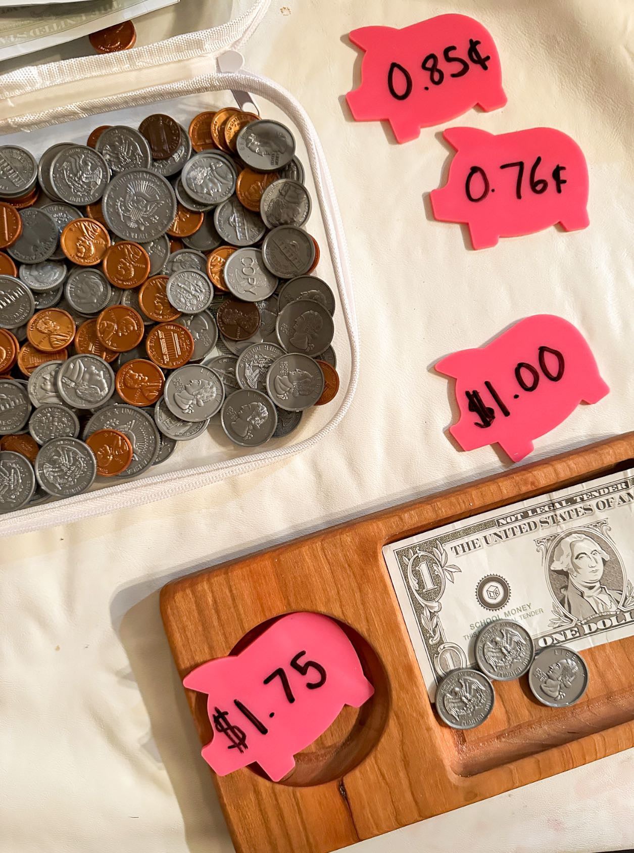 Assorted U.S. coins inside a white pouch next to pink pig-shaped price tags and wooden tray holding a dollar bill and coins