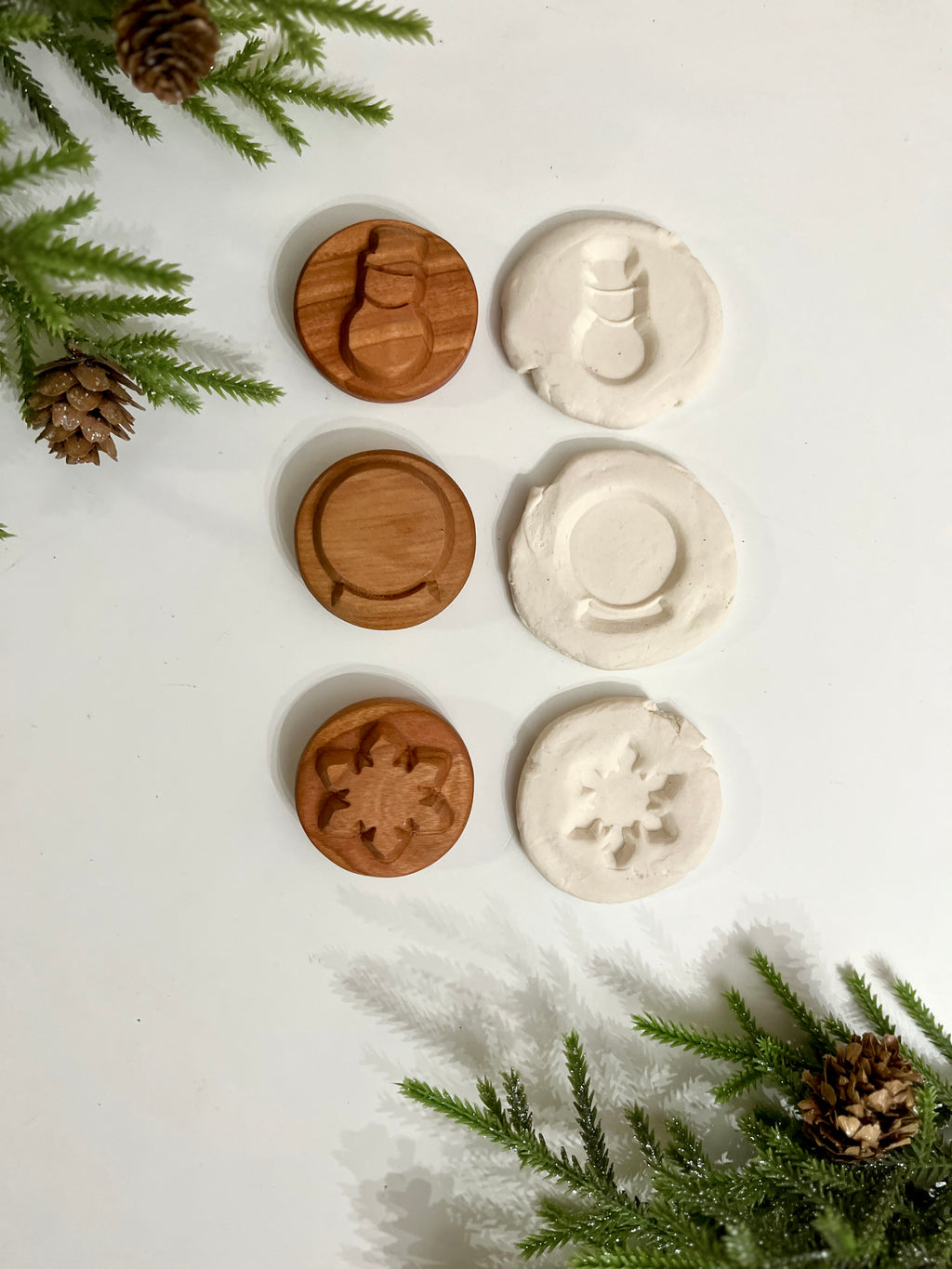 Three wooden stamps with holiday shapes and their white clay imprints surrounded by pine branches and cones on white surface