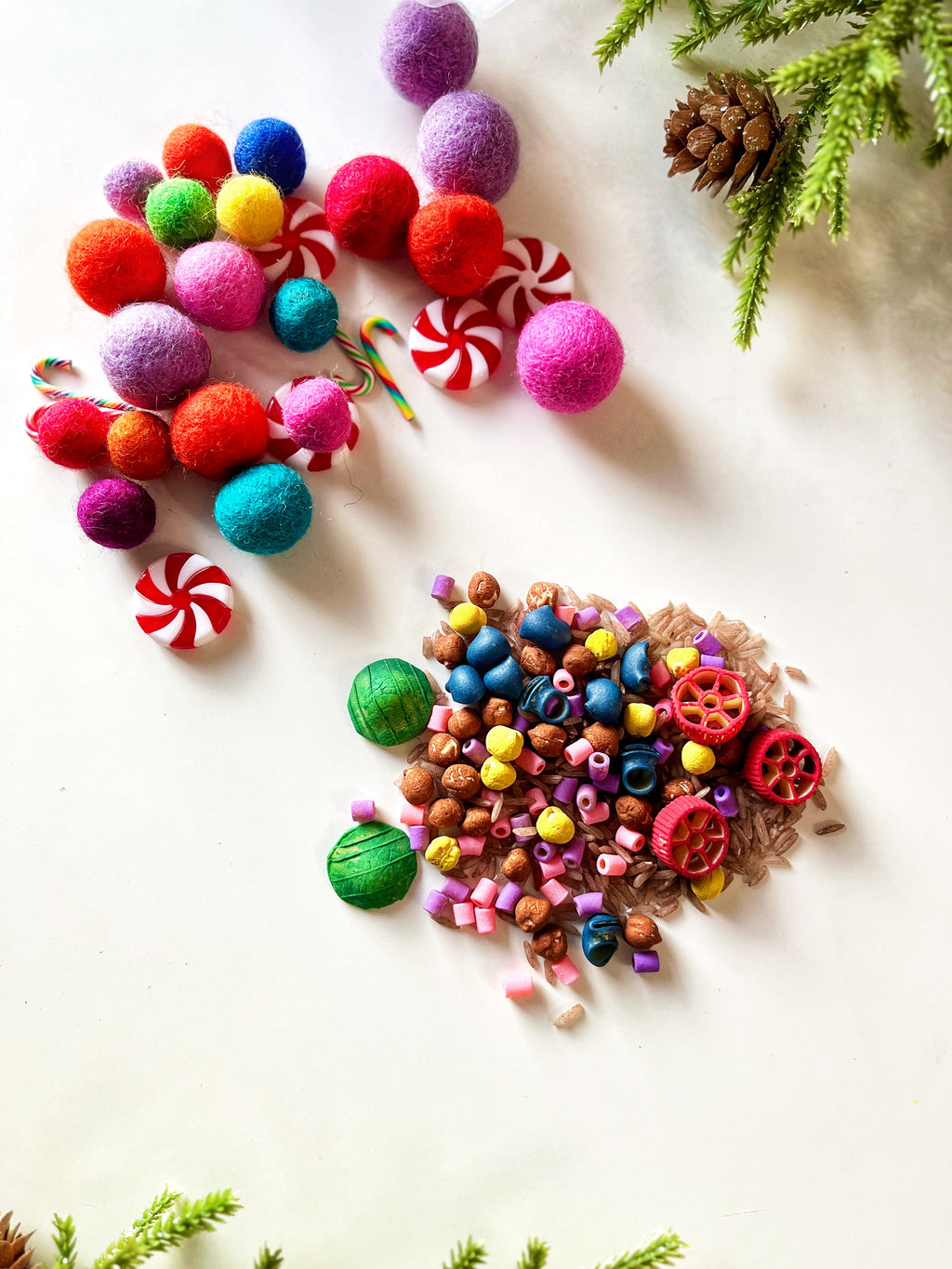 Colorful felt balls, peppermint candy charms, and various small beads scattered on white surface with pine branches and cone accents.