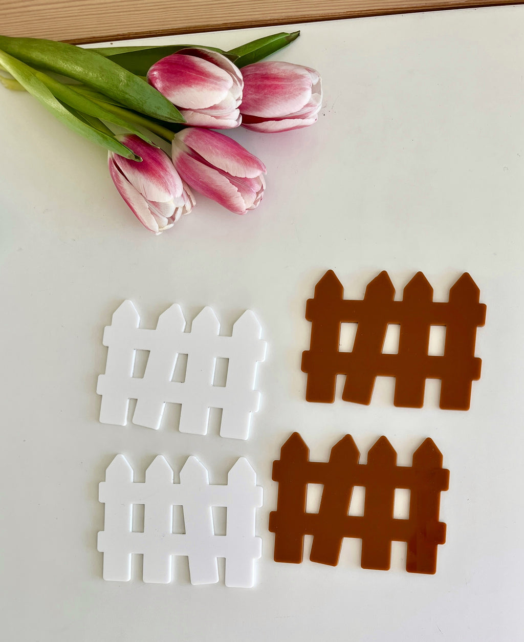 Two white and two brown plastic fence cutouts displayed beside three pink and white tulips on a white surface