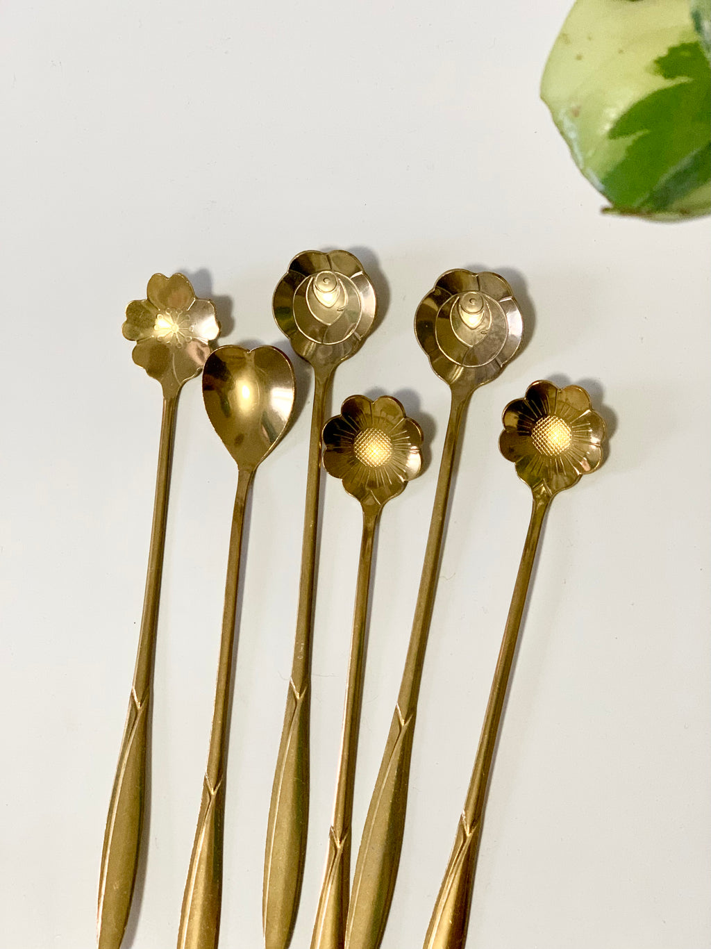 Set of six golden spoons with assorted flower and heart-shaped bowls arranged on white surface with a variegated green leaf nearby