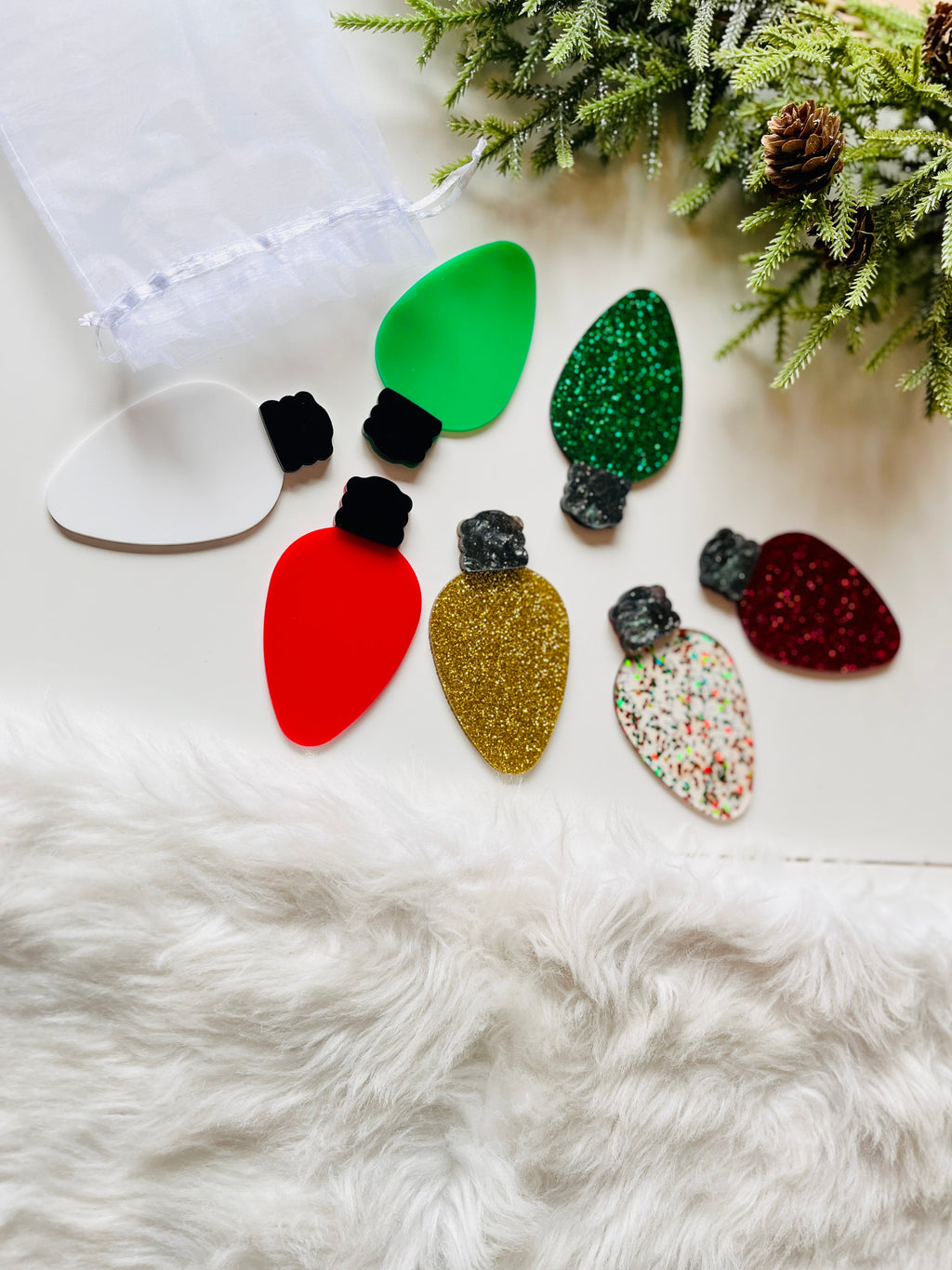 Assorted colorful acrylic Christmas light bulb shapes on white surface with greenery and white fur accents nearby