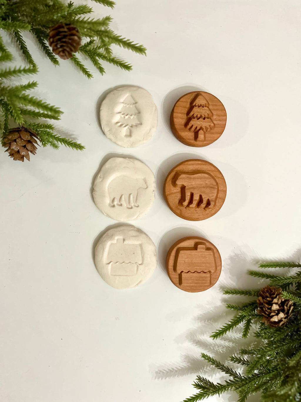 Three wooden stamps with tree, bear, and house shapes alongside matching white clay imprints on white surface with pine branches and cones