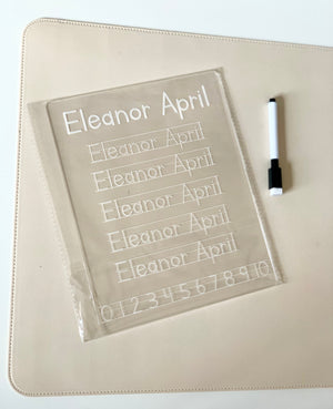 Clear plastic handwriting practice board with the name Eleanor April and numbers alongside a black and white marker on a beige mat