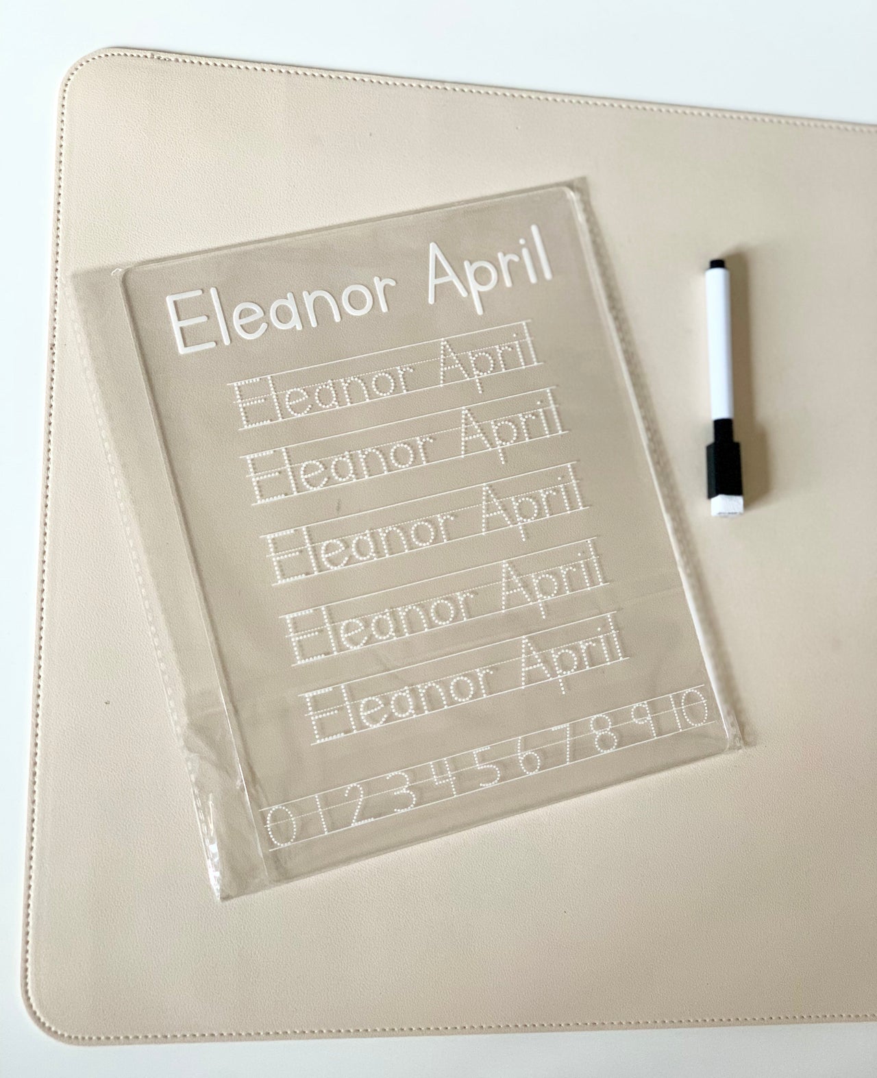 Clear plastic handwriting practice board with the name Eleanor April and numbers alongside a black and white marker on a beige mat