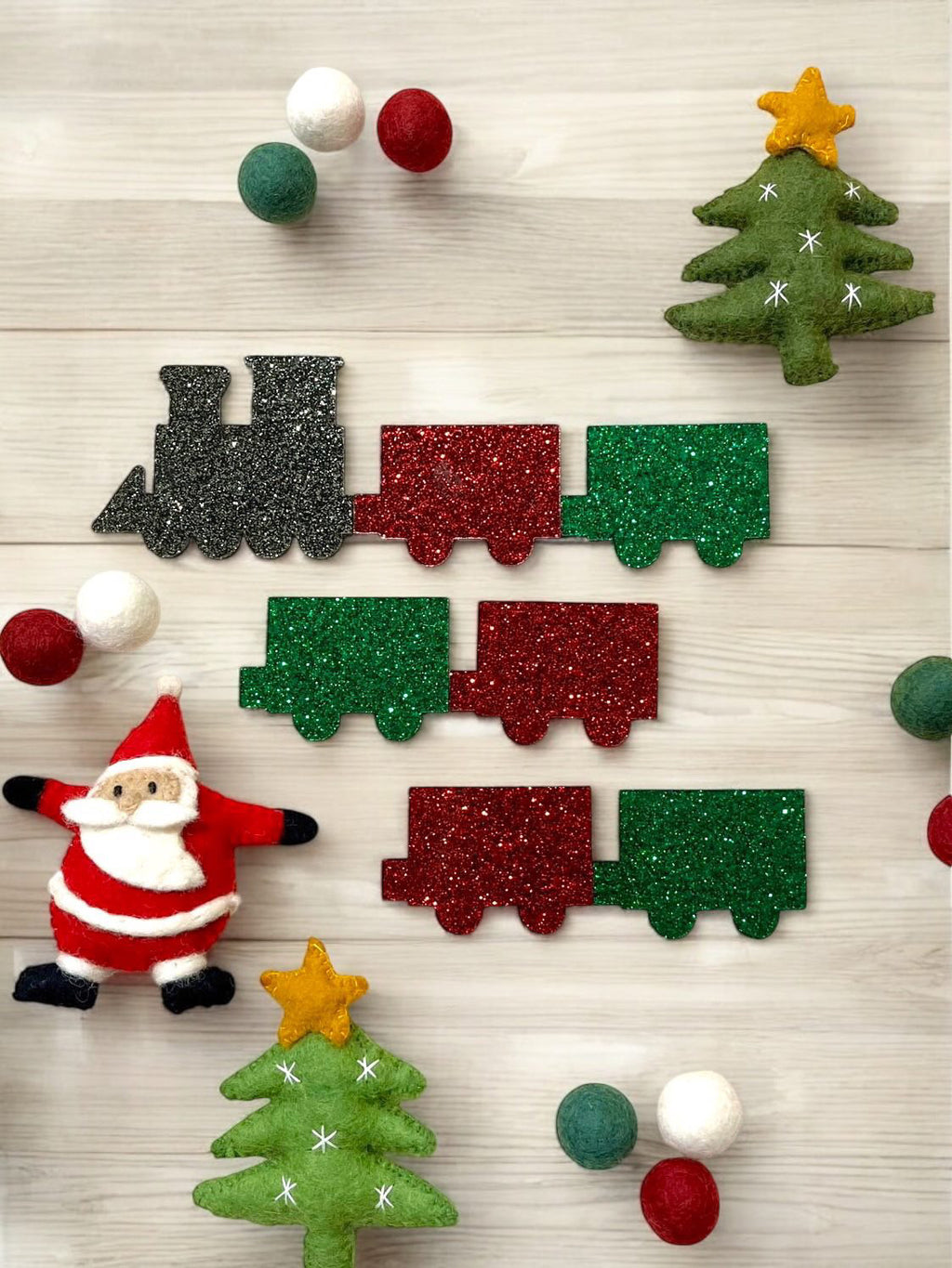 Glittery toy train with red, green, and black cars surrounded by felt Christmas trees, Santa figure, and colored felt balls on wood
