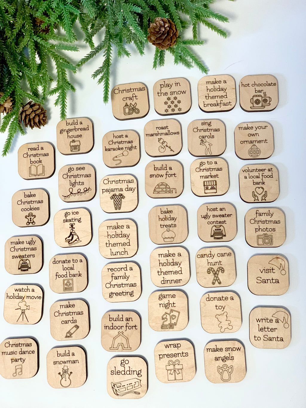 Wooden Christmas activity tokens with festive icons and holiday activity prompts arranged on white surface with evergreen branches and pine cones