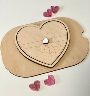 Light wooden heart-shaped box with engraved ribbon design and small white heart knob, surrounded by pink glitter hearts