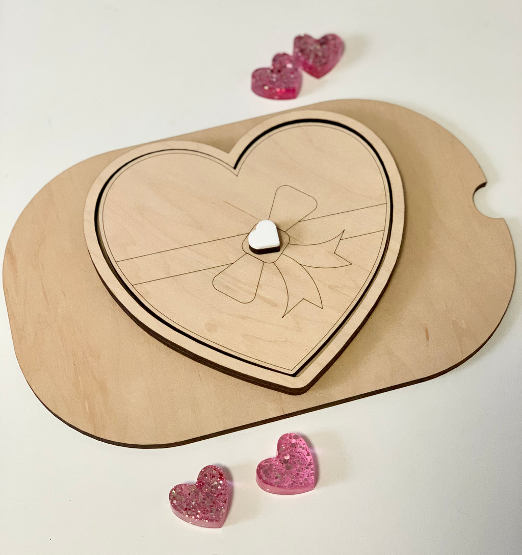 Light wooden heart-shaped box with engraved ribbon design and small white heart knob, surrounded by pink glitter hearts