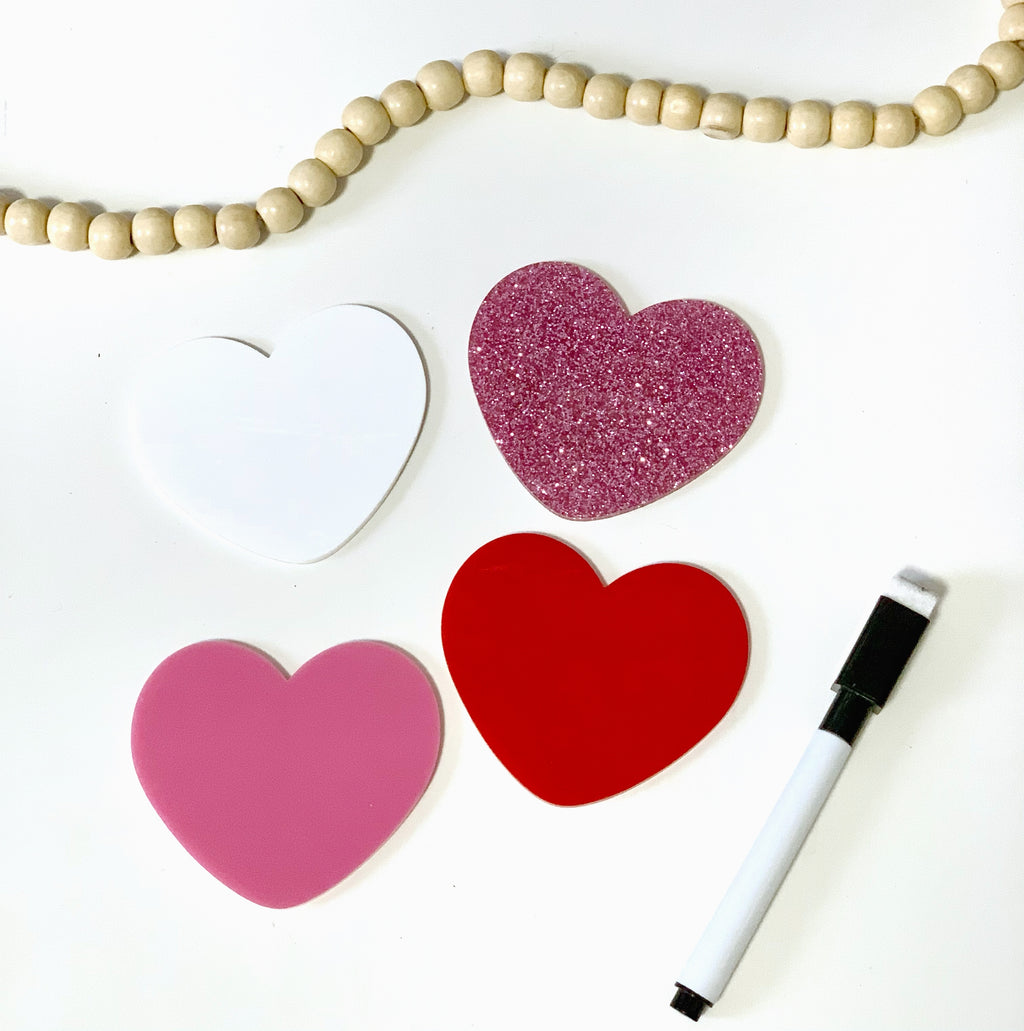 Four heart-shaped boards in white, pink, red, and glitter pink arranged with a dry erase marker and wooden beads above