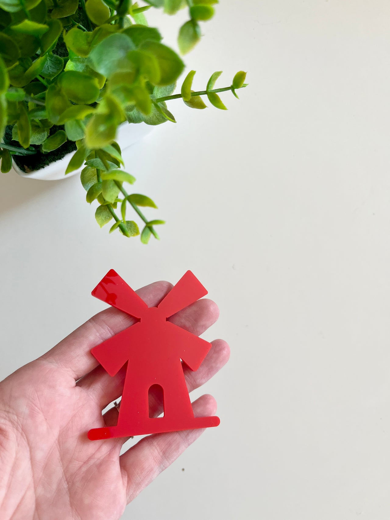 Hand holding a glossy red windmill-shaped cutout with green leaves from a potted plant above on a light surface