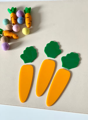 Three flat yellow carrot-shaped cutouts with green tops on a beige surface near small colorful felt balls and fabric carrots