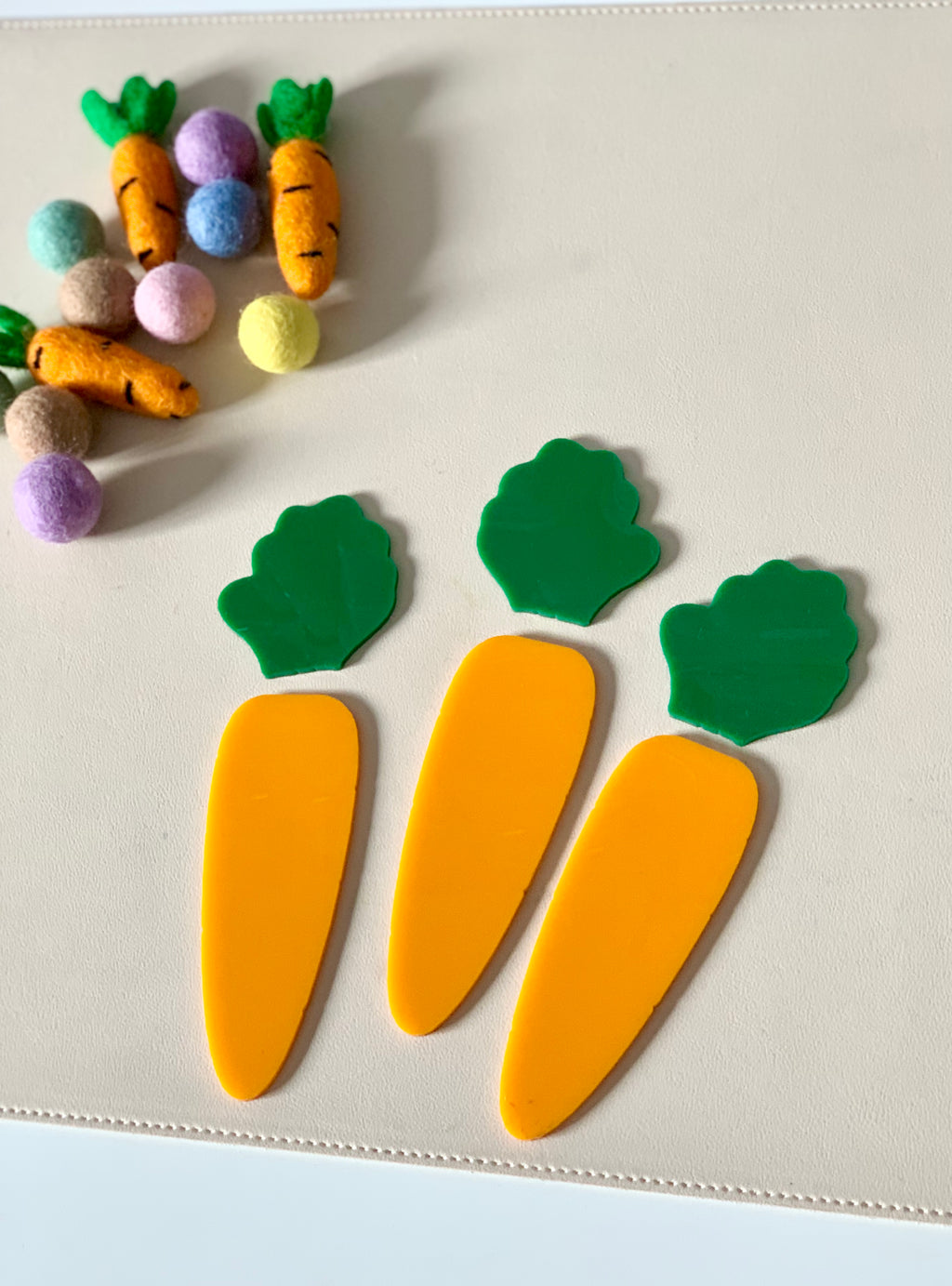 Three flat yellow carrot-shaped cutouts with green tops on a beige surface near small colorful felt balls and fabric carrots