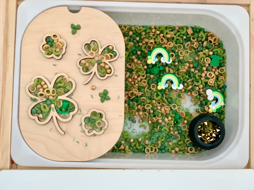 Sensory bin with green and natural colored pasta, wooden shamrock shapes, small beads, rainbows, and a black pot with gold pieces