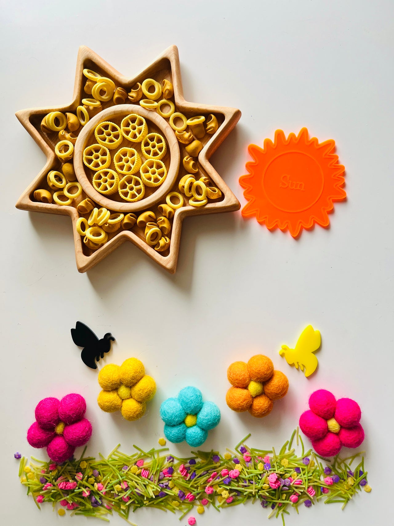 Wooden star-shaped tray with yellow pasta wheels, orange sun-shaped cutter, colorful felt flowers, bees, and green floral grass arrangement on white background