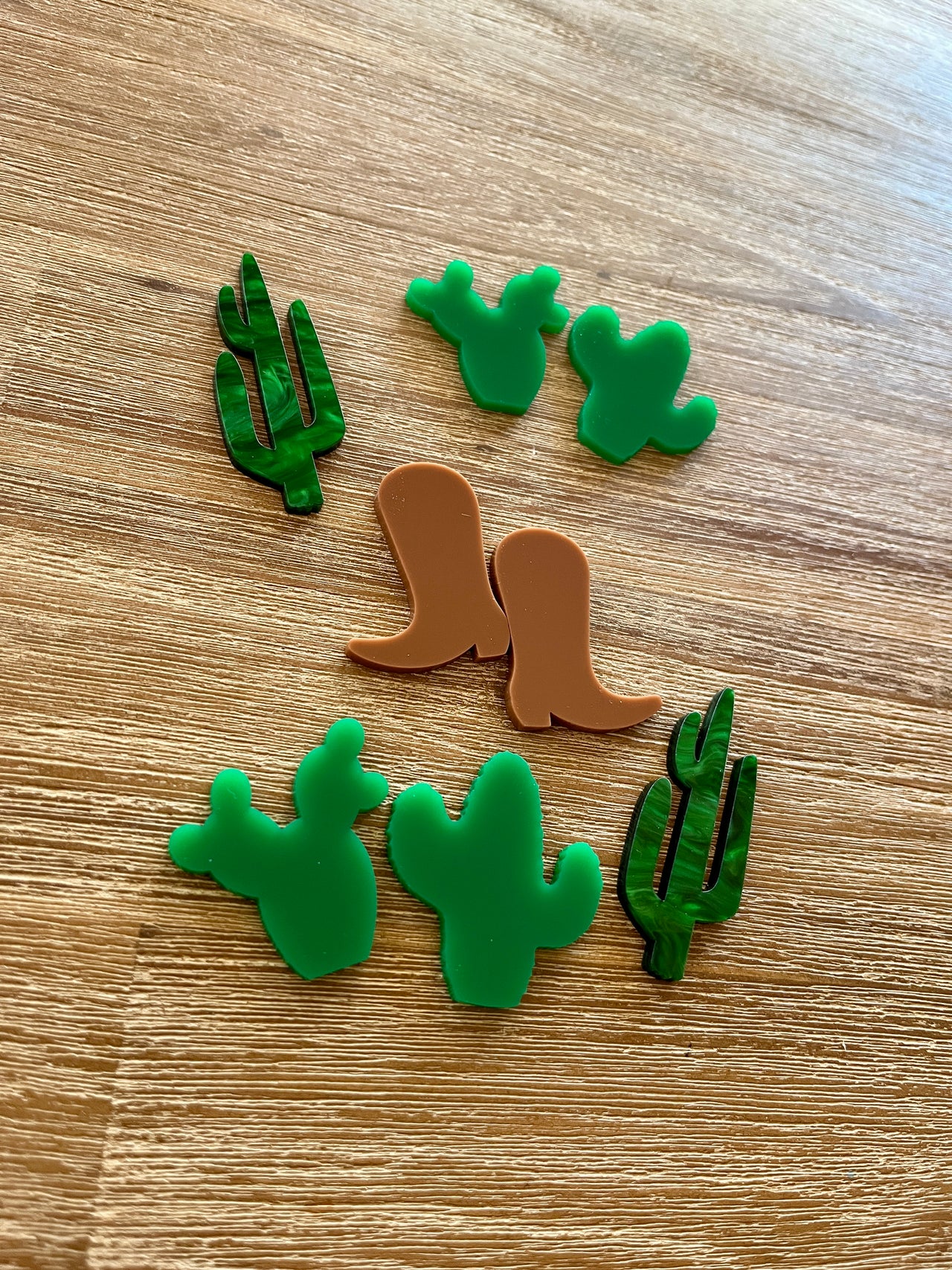 Seven small flat shapes made of durable material arranged on a wooden surface including two translucent green cacti, four solid green cacti, and a pair of brown boots, Little Dry Erasables - Cacti & Boots, handcrafted, homeschool, education resources.