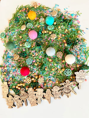 Colorful sensory mix with green pine needle pieces, felt balls, gold beads, wooden snowflakes, and ballet-themed figurines on white surface