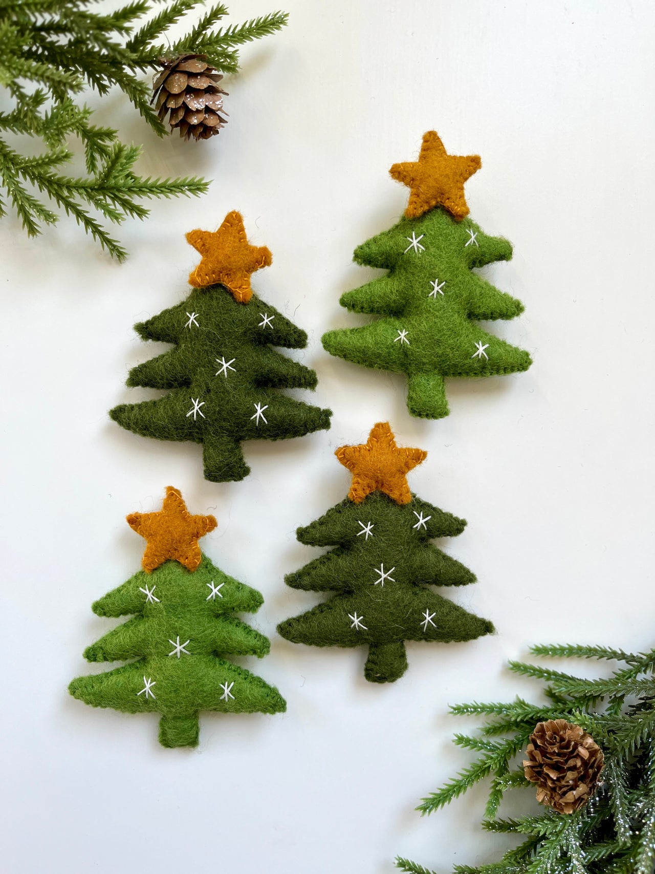 Four felt Christmas tree ornaments with orange stars and white stitched snowflakes on white background with pine branches and cones