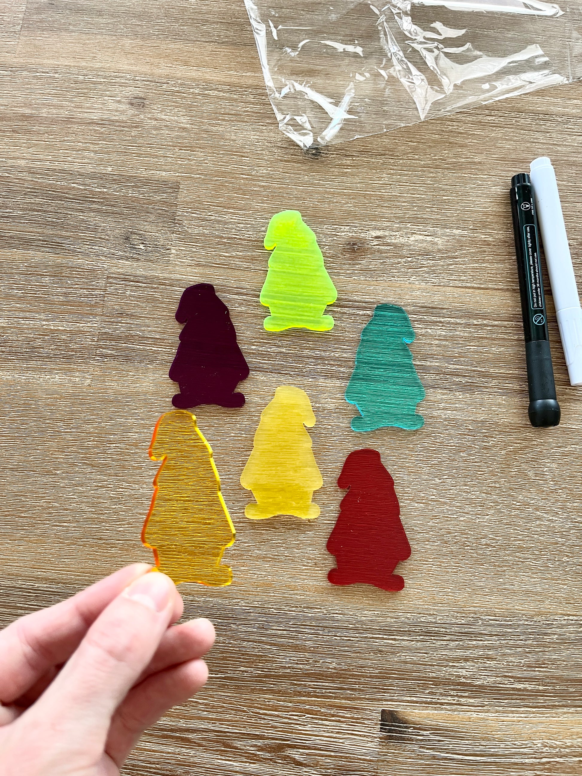 Hand holding a translucent yellow gnome-shaped piece with five additional colorful translucent gnomes and black and white dry erase markers on a wood surface, featuring Little Dry Erasables - Translucent Gnomes, handcrafted homeschool education resources.