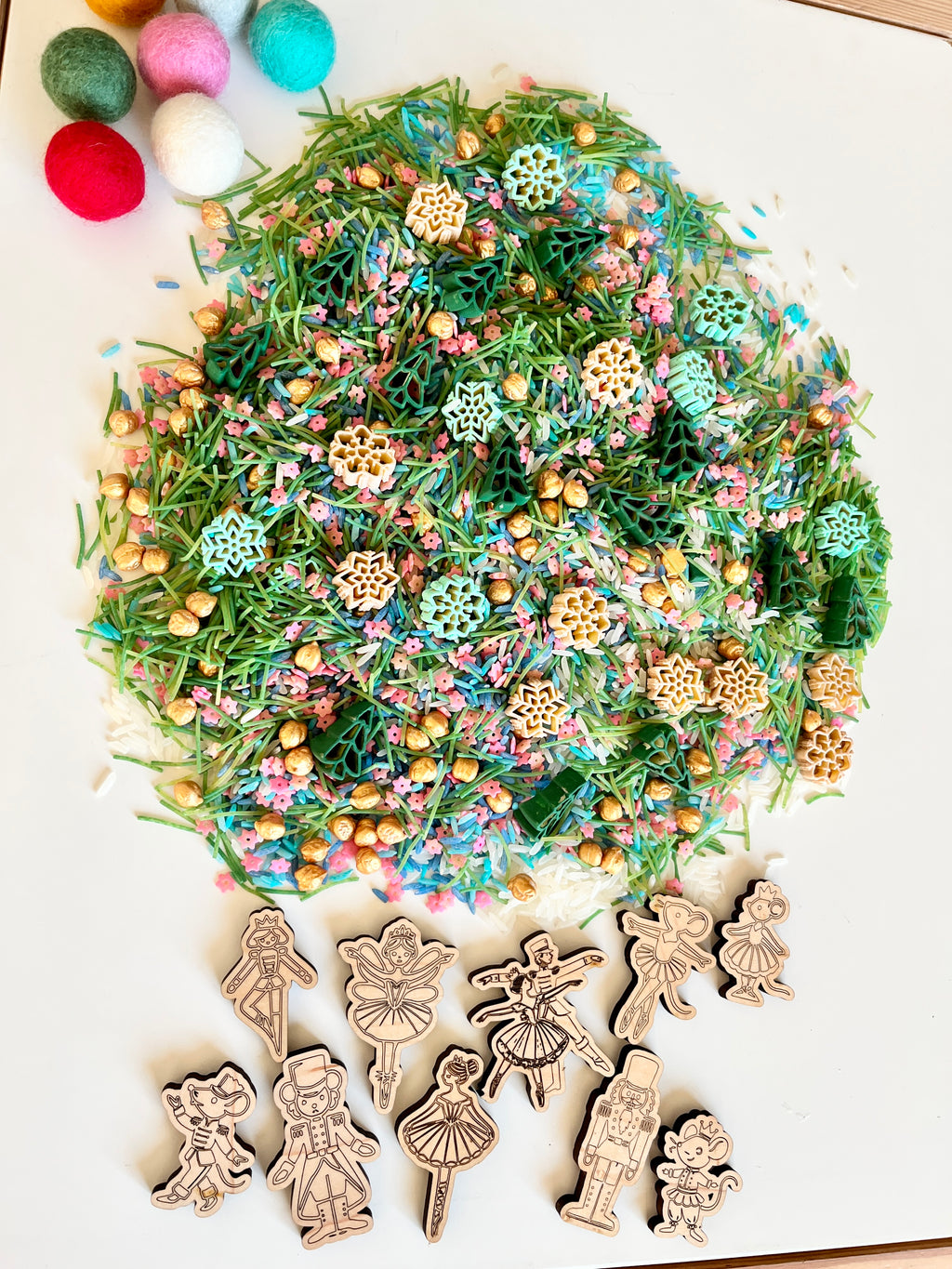Pile of green pasta shaped like trees and snowflakes with colorful sprinkles and wooden ballet-themed character cutouts on white surface