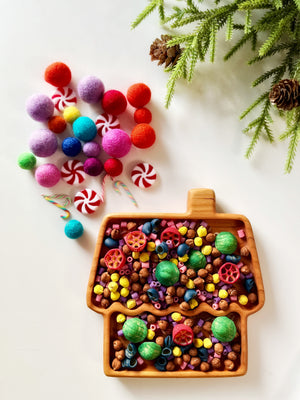Wooden house-shaped tray filled with colorful beads and decorations, surrounded by felt balls and greenery with pine cones