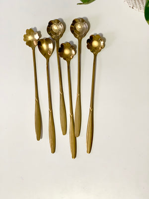 Six long brass spoons with decorative flower-shaped bowls arranged on a white surface with green leaves nearby