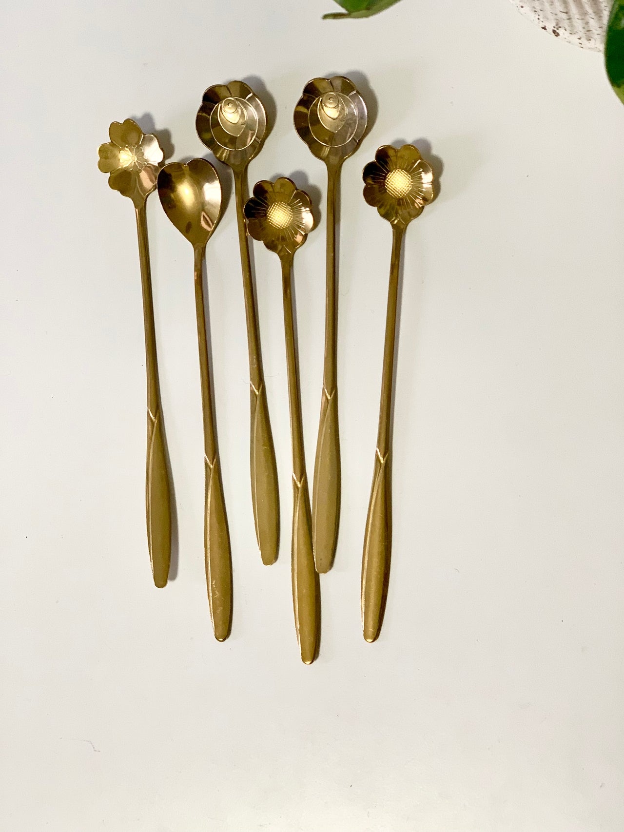 Six long brass spoons with decorative flower-shaped bowls arranged on a white surface with green leaves nearby