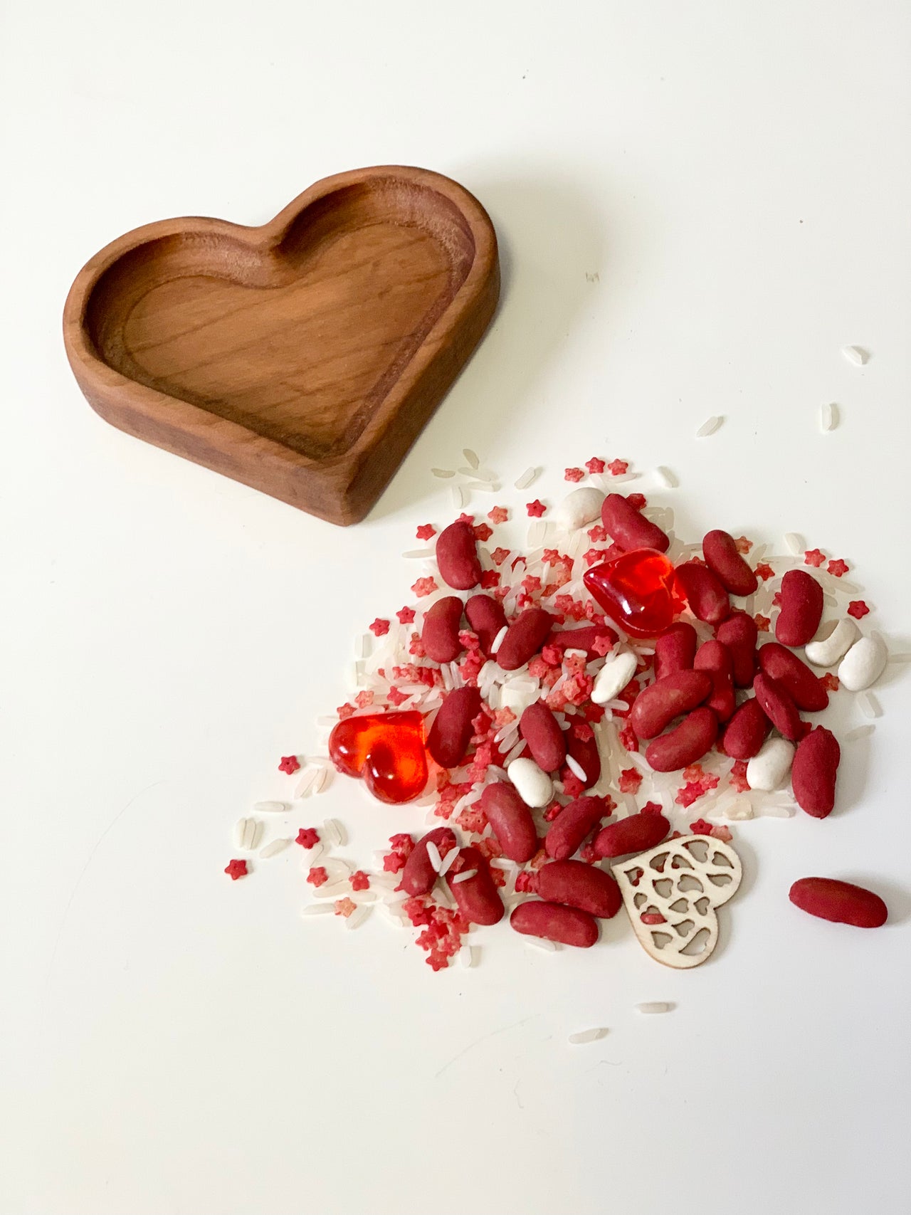 Empty wooden heart-shaped dish next to scattered white rice, red beans, red star-shaped sprinkles, and heart decorations