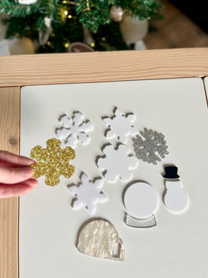 Hand holding gold glitter snowflake among white and silver acrylic holiday shapes including snowman, snow globe, and igloo on table