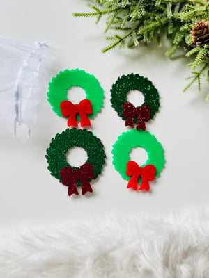 Four small green Christmas wreath cutouts with red bows, two glittery and two solid, on white surface with greenery nearby