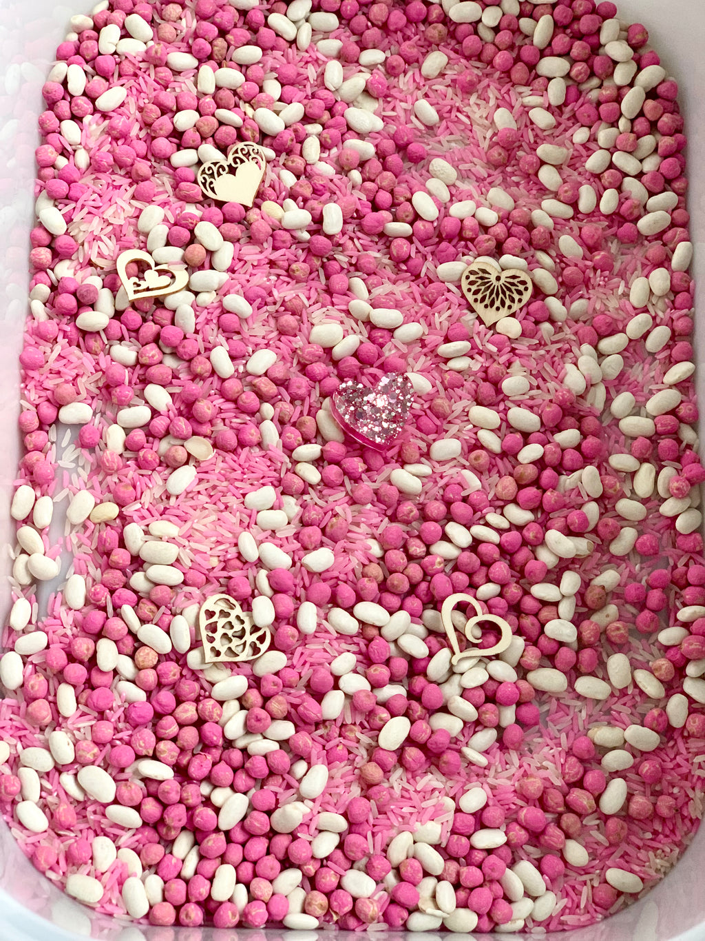 Pink and white sensory bin filled with dyed rice, beans, pink beads, and scattered wooden heart-shaped decorations
