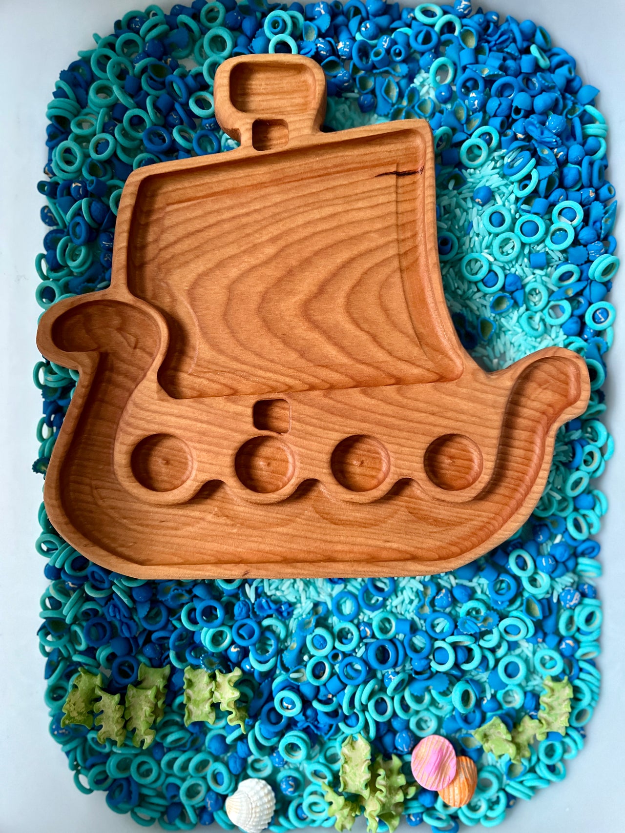 Wooden Viking ship-shaped tray with carved compartments resting on blue and green sensory materials with shells at the bottom