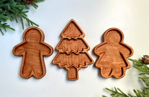 Three smooth wooden trays carved into a Christmas tree and gingerbread man and woman shapes with pine sprigs around