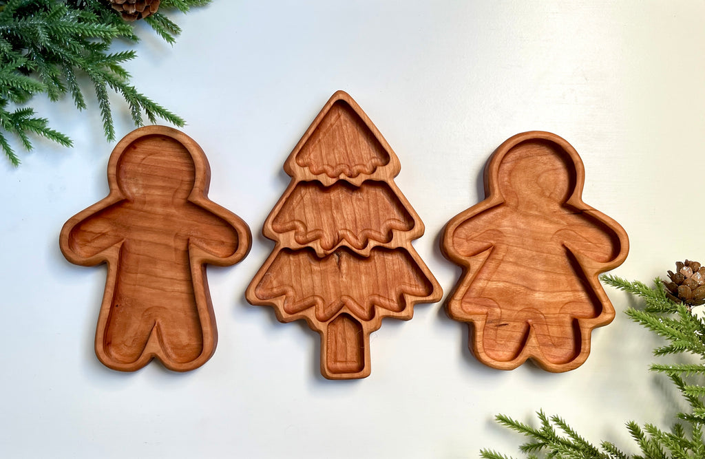 Three smooth wooden trays carved into a Christmas tree and gingerbread man and woman shapes with pine sprigs around