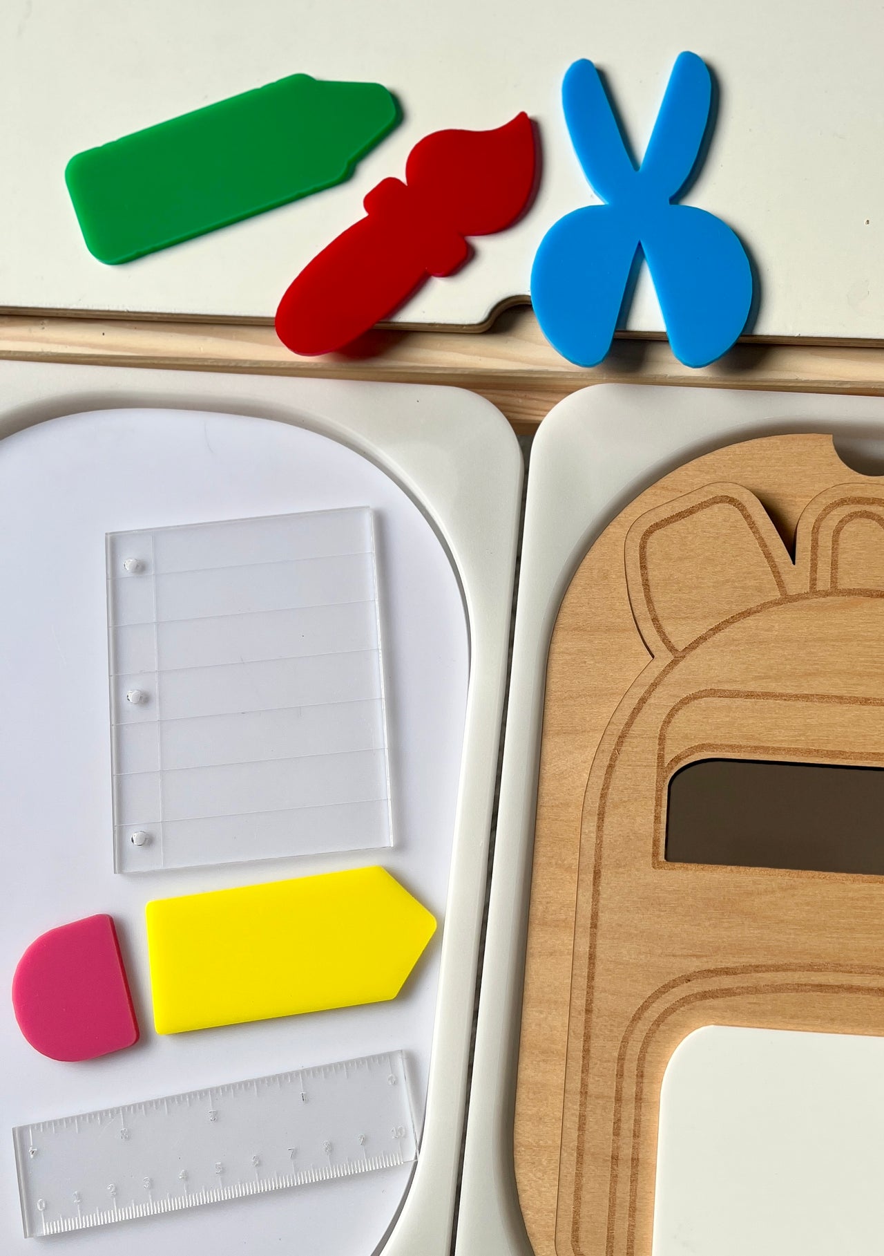 Colorful plastic shapes and clear plastic sheet on white trays beside a wooden puzzle piece with engraved details