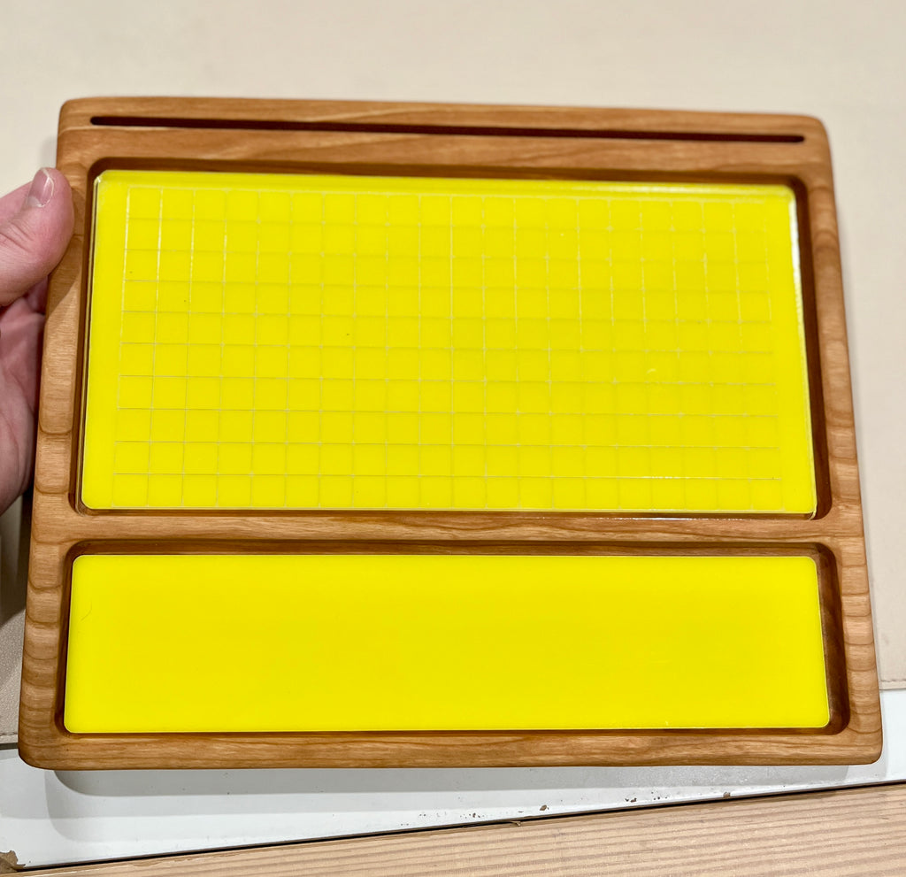 Hand holding a wooden tray with two yellow silicone inserts, one grid-patterned and one smooth, on a beige surface