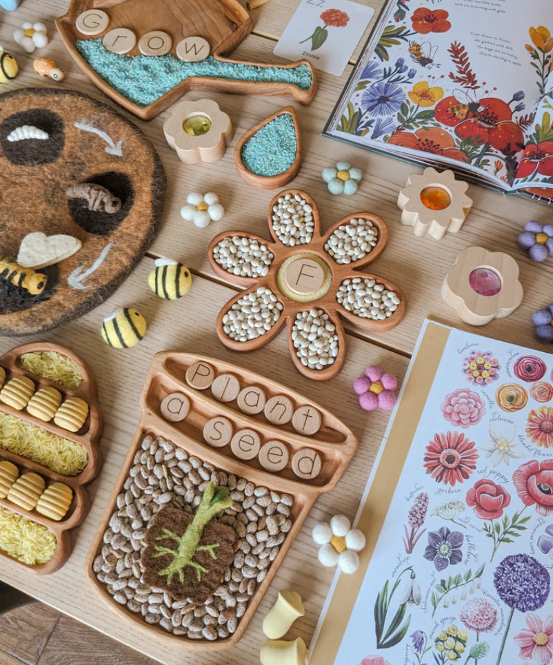 Wooden flower pot plate sensory tray with bean seeds, felt bees, flower-shaped spice jars, and plant-themed books on table