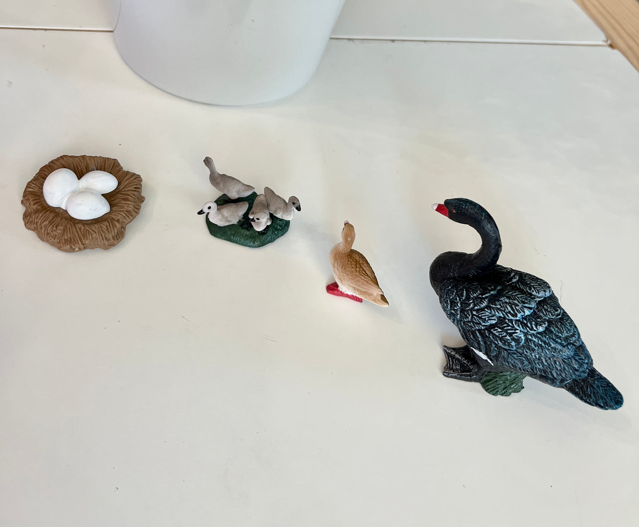 Miniature bird family figurines including white eggs in a nest, ducklings on grass, a small duck, and a black swan on white surface