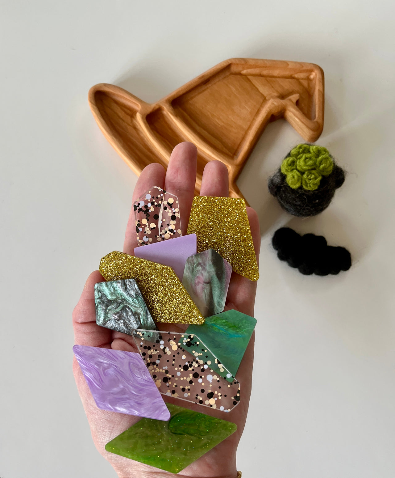 Hand holding assorted geometric acrylic shapes in green, purple, gold glitter, marble, and confetti patterns with wooden tray and felted decorations nearby