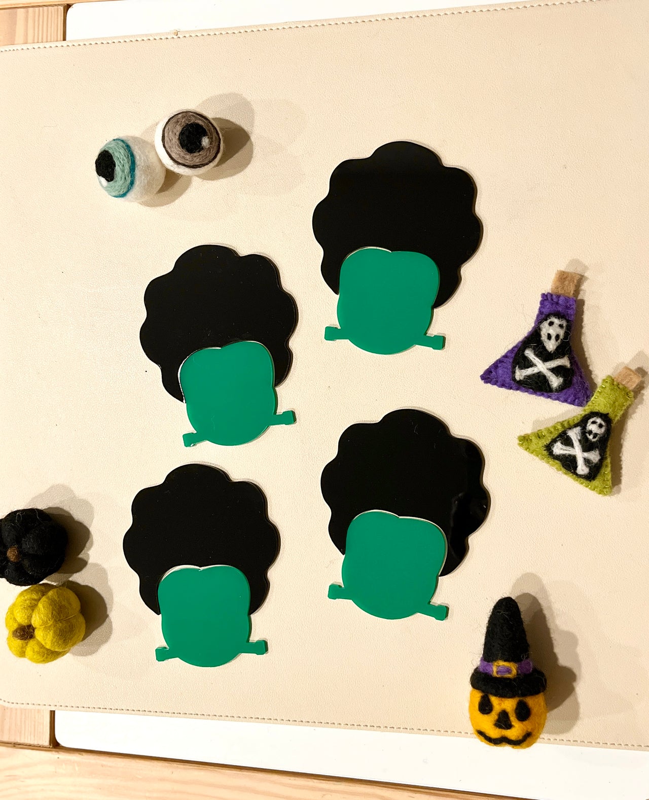 Four Frankenstein-shaped magnets with green faces and black hair surrounded by small Halloween-themed felt decorations on a beige surface