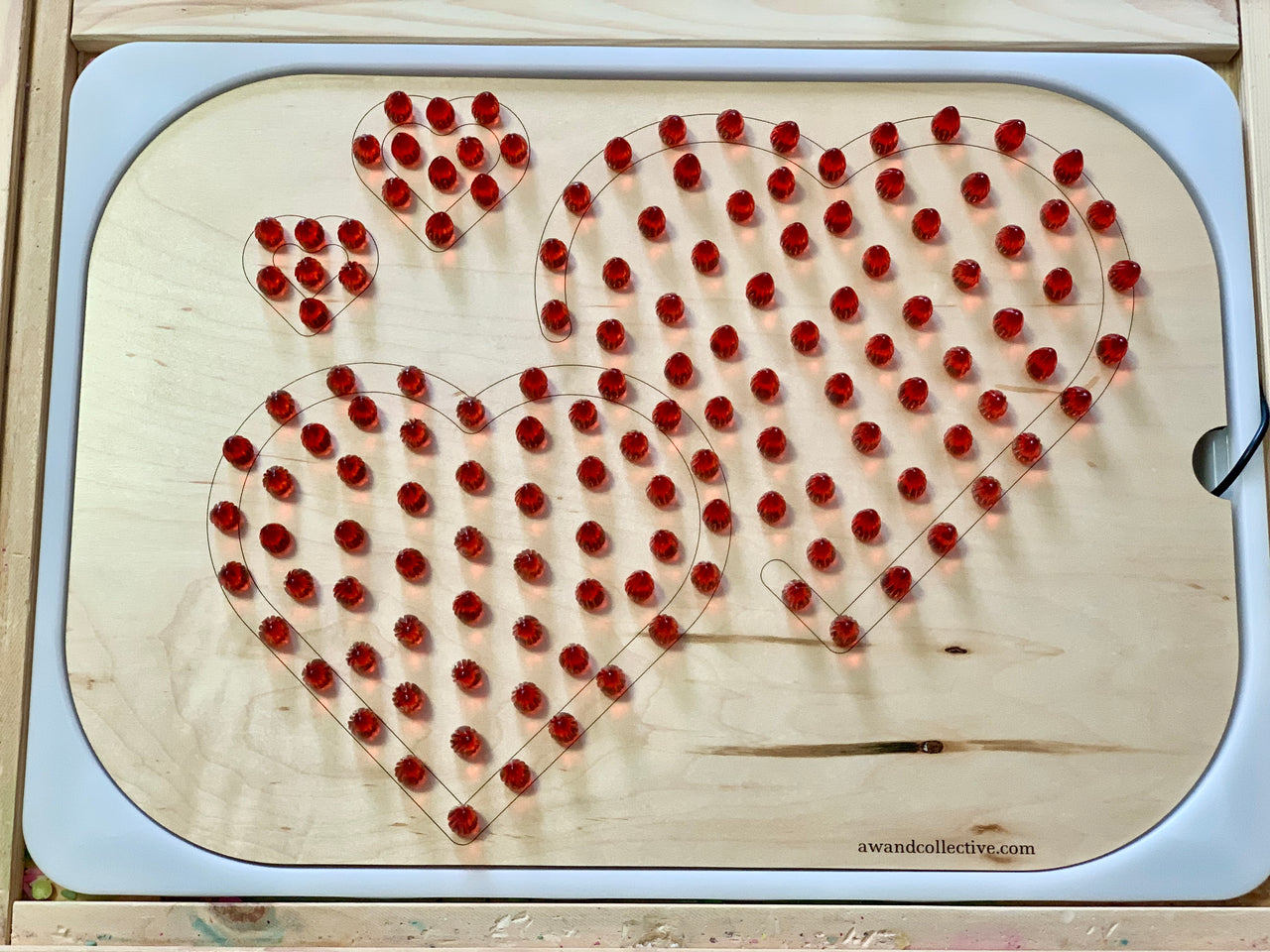 Wooden board with four heart outlines filled with removable red pegs on a light wood background.
